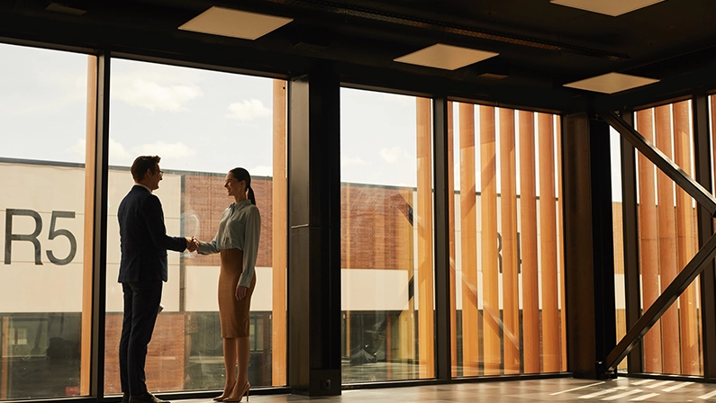 Wide angle view of real estate agent shaking hands with client while standing in empty office building interior lit by sunlight, copy space