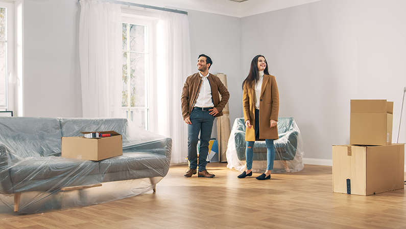 Happy and Excited Young Couple Look Around In Wonder at their Newly Purchased / Rented Apartment. Beautiful People Poses Happily. Big Bright Modern Home with Cardboard Boxes Ready to Unpack.