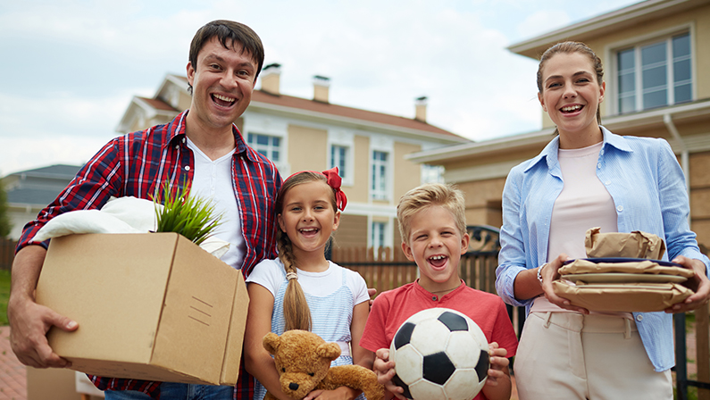 Portrait of happy family with two children, boy and girl, standing holding cardboard boxes and personal belongings outside, in front of their new house, smiling brightly, ready to move in