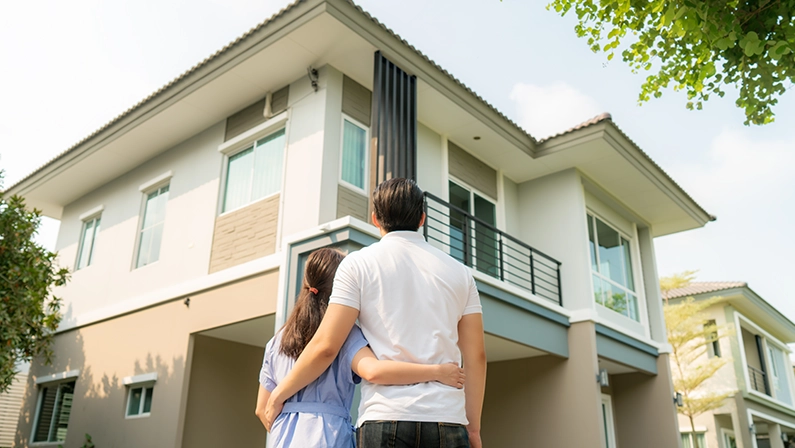 Back portrait of Asian young couple standing and hugging together looking happy in front of their new house to start new life. Family, age, home, real estate and people concept.