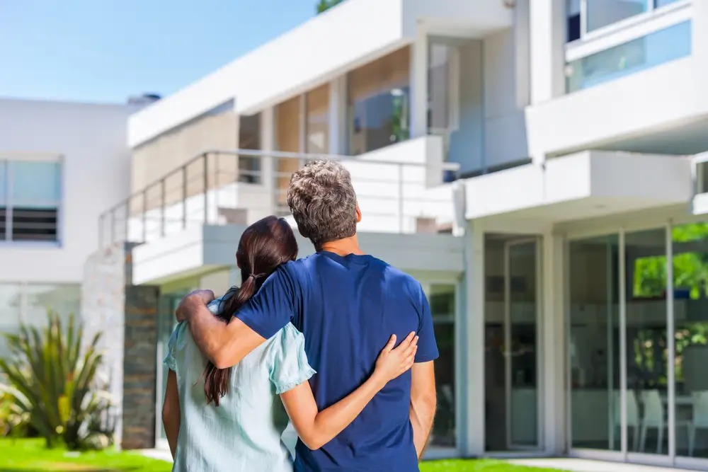 couple embracing in front of new big modern house, outdoor rear view back looking at their dream home