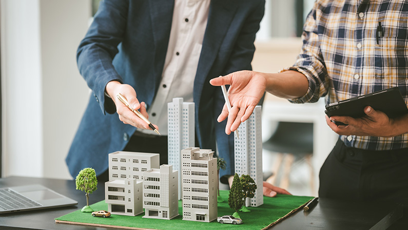 Two middle-aged Asian people businessmen, one holding wind turbine model, discussing over model city, likely about clean energy and new housing projects.